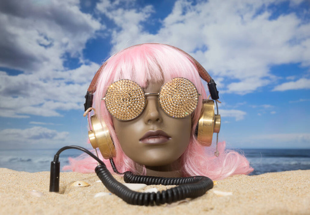 Une tête de mannequin femelle en plastique coincée dans le sable avec des lunettes de soleil scintillantes et des écouteurs sur une plage - Photo, image