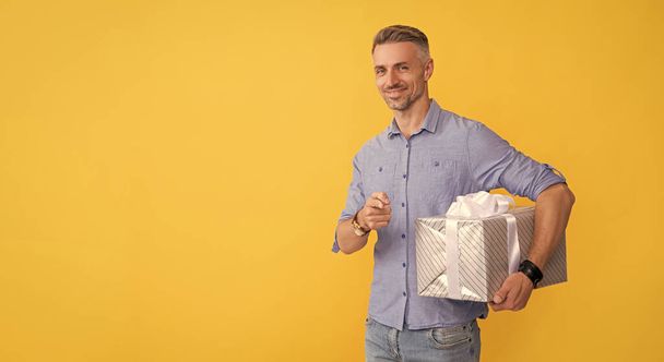 gelukkig volwassen man hold box wijzend vinger op gele achtergrond, kopieerruimte, winkelen. - Foto, afbeelding