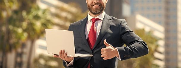 przycięte brodaty biznesmen w formalwear trzymać komputer poza biurem pokazuje kciuk w górę, networking. - Zdjęcie, obraz