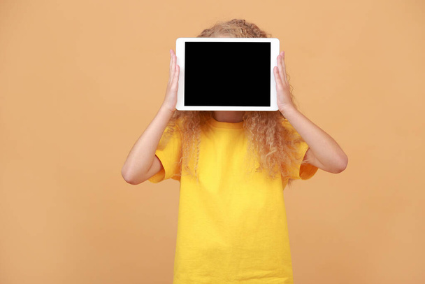 Niesamowity dzieciak ukrywający się za cyfrowym tabletem z pustym ekranem na żółtym tle studia. Podekscytowana uczennica pokazująca blok, makietę, reklamę słodkich aplikacji. Nowa aplikacja edukacyjna, strona internetowa. - Zdjęcie, obraz