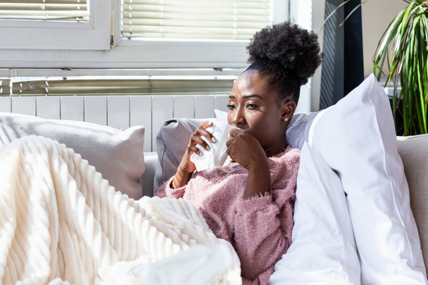 Sick desperate woman has flu. Rhinitis, cold, sickness, allergy concept. Pretty sick woman has runnning nose, rubs nose with handkerchief and drinking hot beverage. African American woman Sneezing in a tissue - Foto, imagen