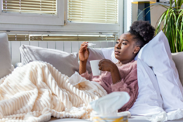Sickness, seasonal virus problem concept. Woman being sick having flu lying on sofa looking at temperature on thermometer. Sick woman lying in bed with high fever. Cold flu and migraine. - Photo, Image