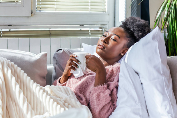 Koud en griep. Portret van een Afro-Amerikaanse vrouw die verkouden werd, zich ziek voelde en niesde in Paper Wipe. Close-up van mooi ongezond meisje bedekt met deken drinken van thee. Concept van de gezondheidszorg. - Foto, afbeelding
