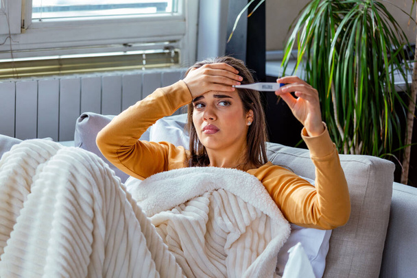 Sickness, seasonal virus problem concept. Woman being sick having flu lying on sofa looking at temperature on thermometer. Sick woman lying in bed with high fever. Cold flu and migraine. - Foto, immagini