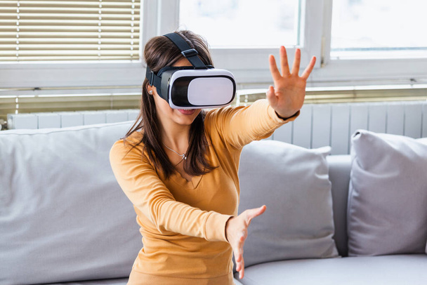 Young elegant woman sitting on sofa chair wearing virtual reality goggles looking online website and using hand moving 3D simulation screen. Enjoying new reality. Attractive young woman in VR headset - 写真・画像