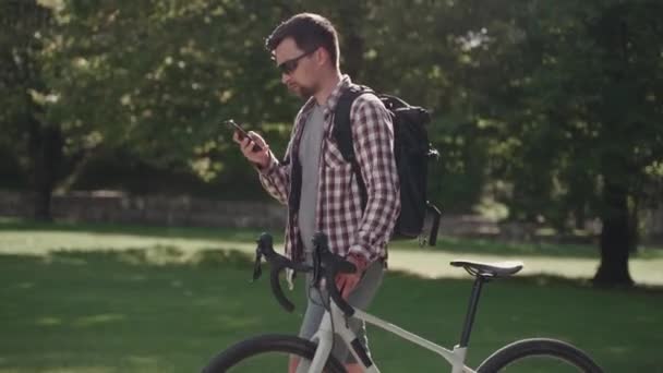 Un ciclista masculino pasea en bicicleta por un parque y usa un teléfono inteligente en un clima soleado en Alemania. Un hombre está empujando una bicicleta y navegando por Internet en su teléfono en un parque de la ciudad en Europa  - Metraje, vídeo
