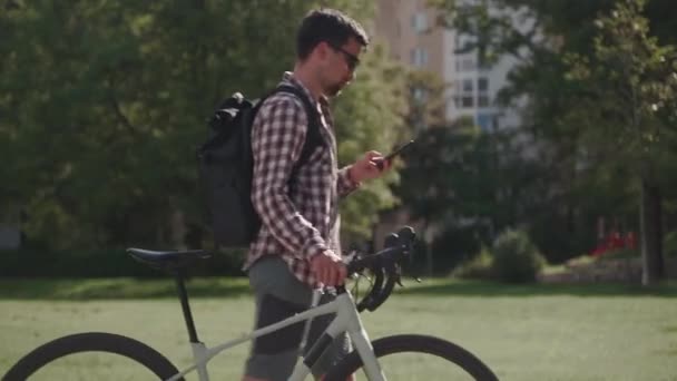 Egy férfi sétál a kerékpárja mellett, és okostelefont használ napos időben Németországban. A fickó tolja a biciklijét és szörfözik az interneten. Beszélgetés biciklizés közben. Modern életmód, kapcsolat.  - Felvétel, videó
