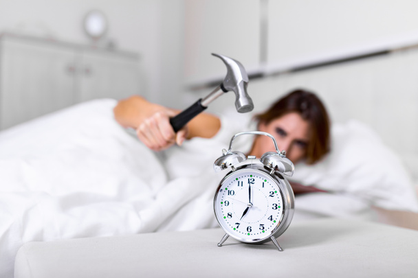 Mujer joven trata de romper el despertador con el martillo, Destruir el reloj. Chica acostada en la cama apagando un despertador con martillo en la mañana a las 7 am. - Foto, Imagen
