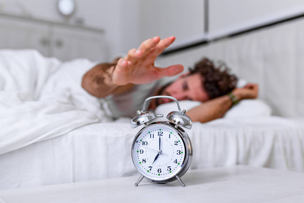 Man lying in bed turning off an alarm clock in the morning at 7am. Hand turns off the alarm clock waking up at morning, man turns off the alarm clock waking up in the morning from a call. - Foto, afbeelding
