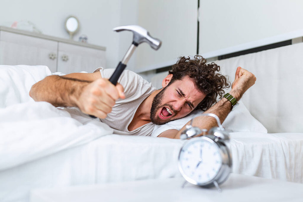 A fiatalember kalapáccsal próbálja eltörni az ébresztőórát, hogy elpusztítsa az órát. Az ágyban fekvő férfi reggel 7-kor kikapcsolja az ébresztőórát kalapáccsal.. - Fotó, kép