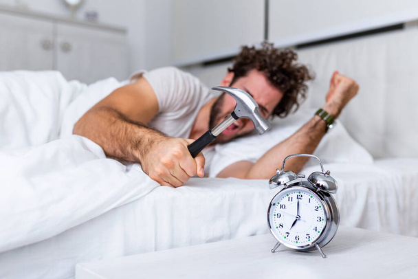 El joven intenta romper el despertador con un martillo, Destruir el reloj. Hombre acostado en la cama apagando un despertador con martillo en la mañana a las 7 am. - Foto, imagen