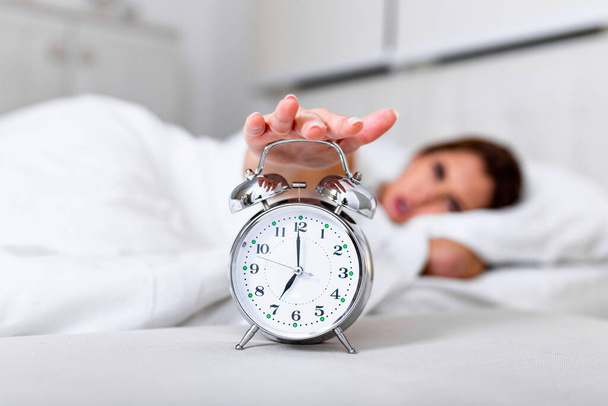 Woman lying in bed turning off an alarm clock in the morning at 7am. Hand turns off the alarm clock waking up at morning, girl turns off the alarm clock waking up in the morning from a call. - Photo, image