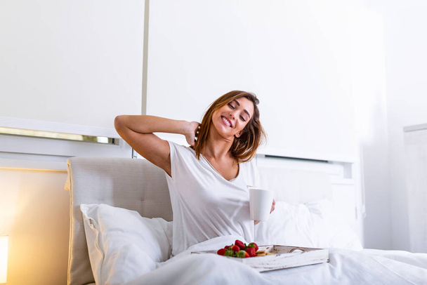 Boldog vonzó nő ébredt fel reggel az ágyban. Nő reggelizik a szállodai szobában, Nő reggelizik az ágyban gyümölcsökkel, kávé és keksz - Fotó, kép