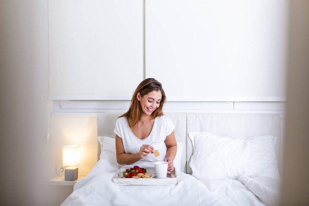 atractiva mujer en pijama despertando por la mañana con el olor de café y desayuno fresco en bandeja de madera en la cama - Foto, imagen