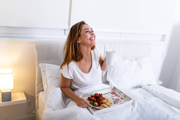 Mosolygó fiatal nő finom reggelit és kávét eszik egy tálcán az ágyban. Egy nő az ágyban reggelizik. Nagy fogú mosoly.. - Fotó, kép