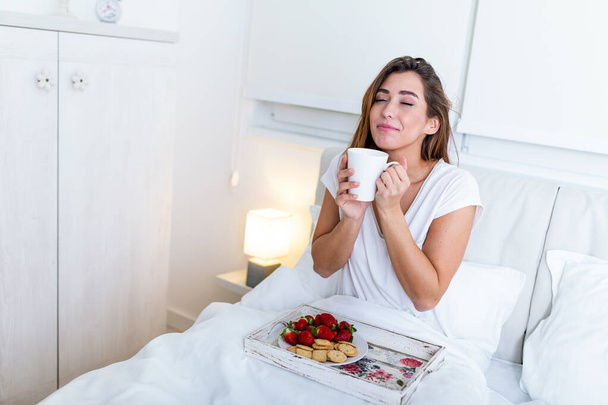 Πρωινό στο κρεβάτι για νεαρή όμορφη γυναίκα. Γυναίκα που πίνει καφέ και πρωινό στο κρεβάτι με φρούτα, καφέ και μπισκότα. Πρωινό ξύπνημα στο ξενοδοχείο - Φωτογραφία, εικόνα