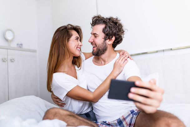 Junges Paar im Bett, Selfie auf dem Handy, junges glückliches schönes Paar auf dem Bett, das Selbstporträt mit dem Smartphone macht und sich umarmt, Morgen-Selfie. Paar im Bett macht süße Fotos - Foto, Bild