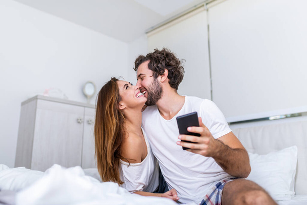 一緒にベッドに横になっているスマート フォン、幸せな彼氏と朝は、若い女性が男に新しい携帯電話アプリケーションを示す社会的なネットワークのニュースをチェックして彼女を使用して魅力的な千年カップルの笑顔 - 写真・画像
