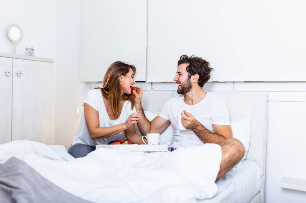 Jonge knappe man voedt aantrekkelijke jonge vrouw in bed in de ochtend. Romantisch ontbijt voor twee. Liefde, zorg, relatioships. koppel gezond ontbijt samen in bed thuis - Foto, afbeelding