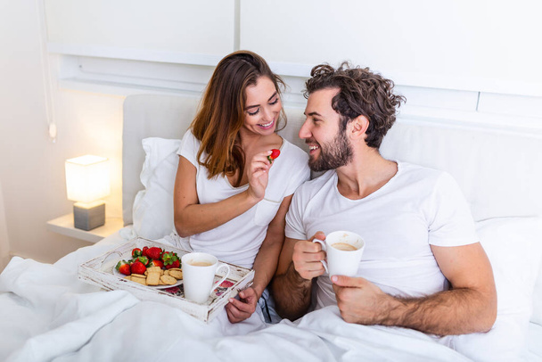 Cute couple having breakfast in bed in the bedroom. Beautiful woman feeding her boyfriend strawberries in bed while having breakfast and coffee in bedroom - Photo, Image