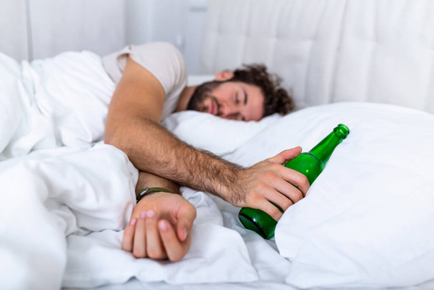 Un ivrogne dans son lit, un endroit triste et une bouteille d'alcool à la main. Jeune homme couché dans son lit ivre mortel tenant une bouteille d'alcool presque vide. - Photo, image