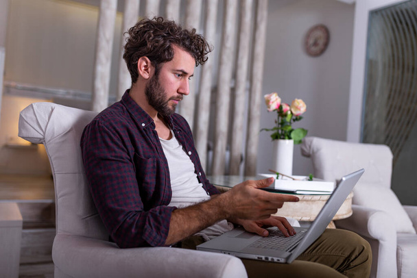 Serious bussines man with serious face sitting at home and using laptop for his project. freelance working from home concept Business man at home checking statistics on mobile phone and laptop - Photo, Image