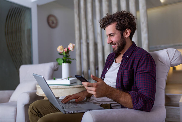 Successful entrepreneur smiling in satisfaction as he checks information on his laptop computer while working in a home office. Young man relaxing on the sofa with a laptop and mobile phone in hands - Φωτογραφία, εικόνα