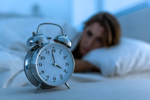 Sleepless and desperate beautiful caucasian woman awake at night not able to sleep, feeling frustrated and worried looking at clock suffering from insomnia in sleep disorder concept. - Фото, изображение