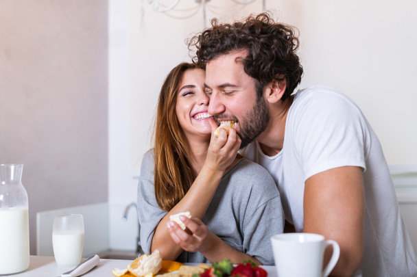 Beautiful young couple is feeding each other and smiling while cooking in kitchen at home. Happy sporty couple is preparing healthy food on light kitchen. Healthy food concept. - Photo, Image