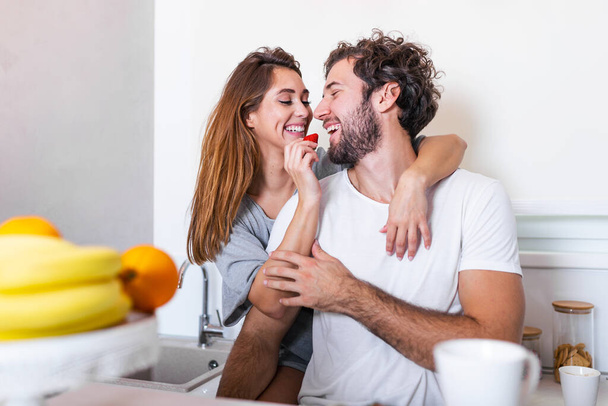 美しい若いカップルは、自宅でキッチンで料理をしている間にお互いを食べ、笑顔です。明るいキッチンで健康的な食べ物を準備している幸せなスポーティカップルです。健康食品のコンセプト. - 写真・画像