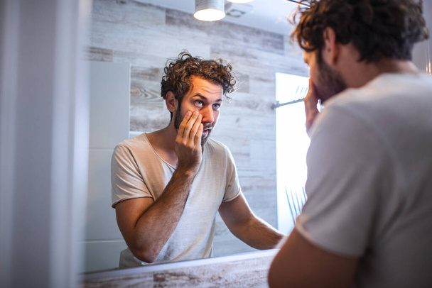 Young caucasian man taking care of his face looking at mirror reflection in bathroom morning clean eyes. Reflection of handsome man with beard looking at mirror and touching face in bathroom grooming - Фото, зображення
