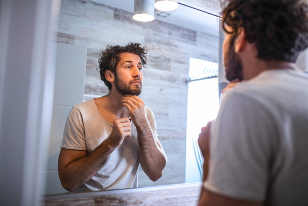 Reflection of handsome man with beard looking at mirror and touching face in bathroom grooming. Man putting skincare facial treatment cream on face. - Fotoğraf, Görsel