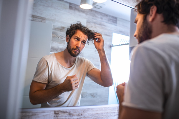 Όμορφος νεαρός άνδρας αγγίζει τα μαλλιά του με το χέρι και περιποίηση στο μπάνιο στο σπίτι. Λευκός μετροσέξουαλ άντρας ανησυχούσε για τριχόπτωση και κοίταζε τον καθρέφτη του.. - Φωτογραφία, εικόνα