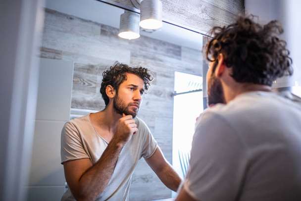 Reggeli higiénia, jóképű férfi a fürdőszobában, tükörbe néz. Tükröződése jóképű férfi szakáll néz tükör és megható arc fürdőszobában ápolás - Fotó, kép