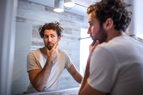Ochtendhygiëne, knappe man in de badkamer die in de spiegel kijkt. Reflectie van knappe man met baard kijken naar spiegel en aanraken gezicht in badkamer verzorging - Foto, afbeelding