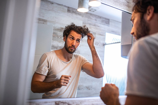 Όμορφος νεαρός άνδρας αγγίζει τα μαλλιά του με το χέρι και περιποίηση στο μπάνιο στο σπίτι. Λευκός μετροσέξουαλ άντρας ανησυχούσε για τριχόπτωση και κοίταζε τον καθρέφτη του.. - Φωτογραφία, εικόνα