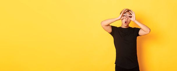 Portrait of funny and cute asian blond guy making fingers glasses and grimacing, standing yellow background. - Photo, Image
