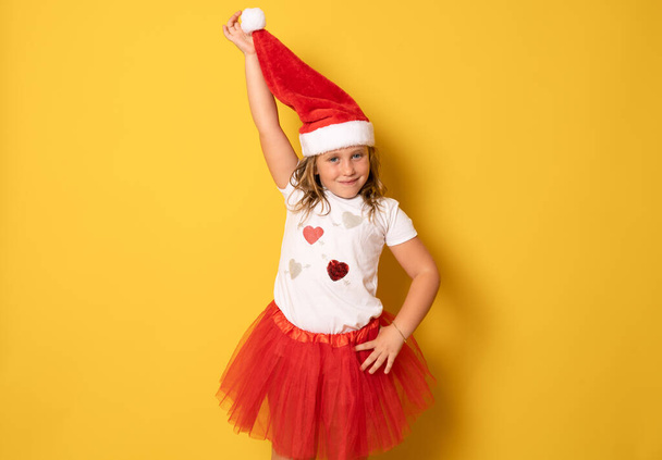 Lustiges nettes kleines Mädchen mit rotem Weihnachtsmannhut, das auf gelbem Hintergrund steht. Porträt - Foto, Bild