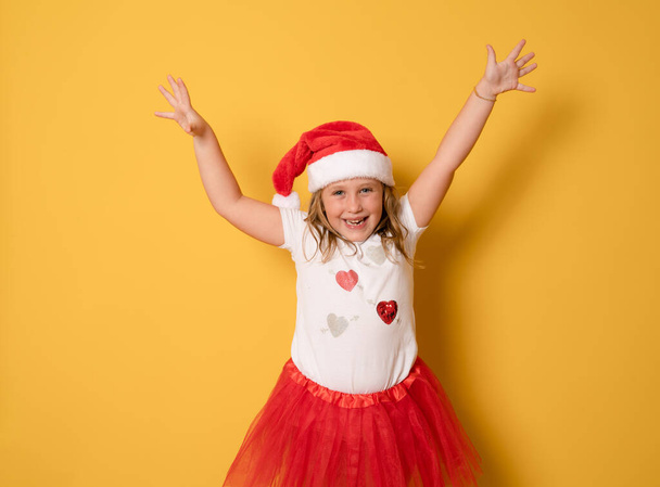 kleines Mädchen mit rotem Weihnachtsmann-Hut und erhobenen Armen auf gelbem Hintergrund. Porträt - Foto, Bild