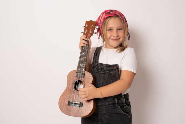 Cute little girl in denim clothing playing ukulele over white background. Happy vacation concept. - Fotografie, Obrázek