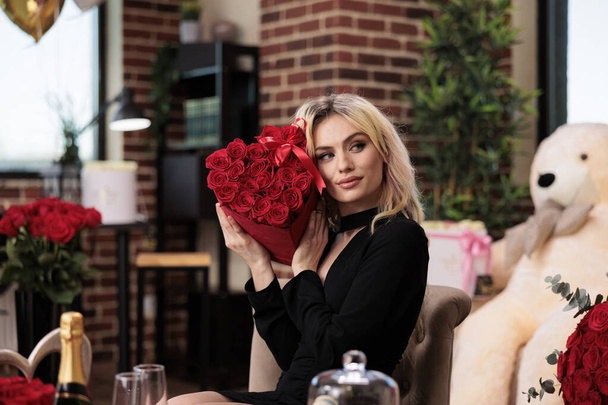 Egy fekete ruhás nő Valentin napi vörös rózsát tart, luxusajándékokat kap. Fiatal szőke lány virágcsokorral ül a nappaliban, tele drága romantikus ajándékokkal - Fotó, kép