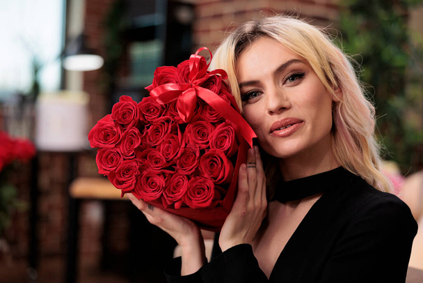 Gyönyörű nő kezében luxus vörös rózsa néz kamera, Valentin napi ajándék. Vonzó szőke barátnő virágcsokor szív alakú doboz portré, romantika ünnep ünnep - Fotó, kép