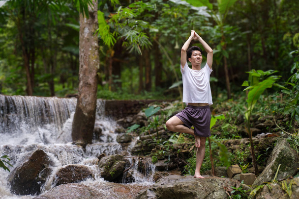 Homme en bonne santé pratiquant le yoga sur les rochers près de la cascade dans la forêt. Concept de calme et de méditation. - Photo, image
