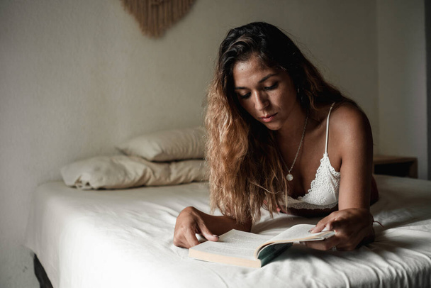 Young Latina girl with long hair reading a book lying on her bed in her room. - Foto, immagini