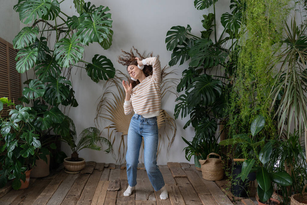 Positiivinen nuori italialainen nainen langattomissa kuulokkeissa kuuntelemassa musiikkia ilolla, nauttimassa hetkestä, rentouttava, tanssia puulattialla viihtyisässä kotipuutarhassa hirviön ja trooppisten kasvien kanssa.  - Valokuva, kuva