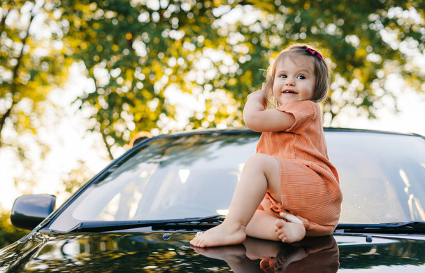 La ragazza che aspetta i suoi genitori, in piedi sul cofano dell'auto. Assistenza all'infanzia. Famiglia felice all'aperto. Stile di vita all'aperto. Famiglia felice, infanzia. Famiglia divertente. - Foto, immagini