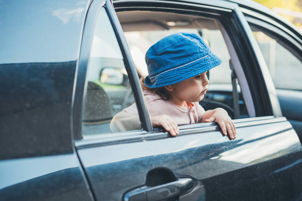 Mała dziewczynka w kapeluszu wystająca głową przez okno samochodu, widząc piękne widoki na przyrodę. Uśmiechnięte szczęśliwe dziecko. Widok z boku. Osoba emocjonalna. - Zdjęcie, obraz