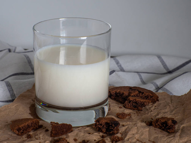 Glass of milk and cookies made from cocoa and with pieces of chocolate inside on a baking paper on a white background. Breakfast eating. Copy space, close up. - Foto, imagen