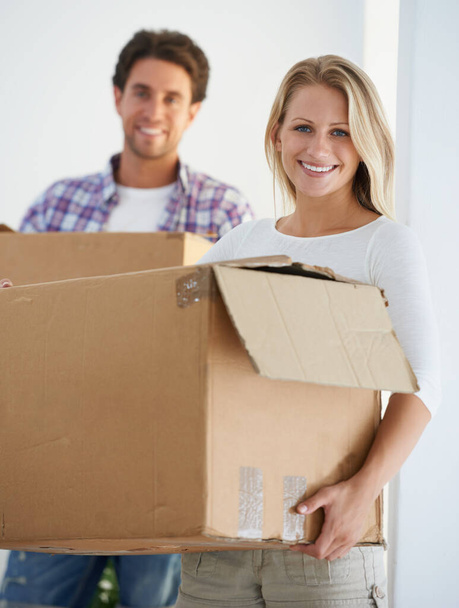 Moving into a home of her own. Portrait of a smiling woman carrying a brown cardboard box with her boyfriend carrying a box in the background - Foto, Bild