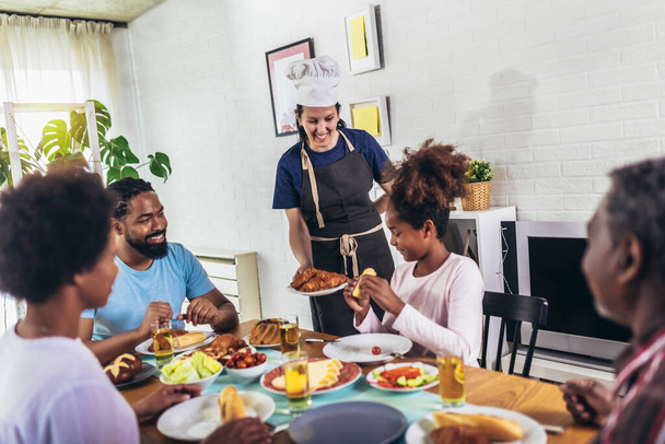 Egy többgenerációs afro-amerikai család, akik a vacsoraasztaluknál esznek. Fehér nő főz szolgálni őket. - Fotó, kép